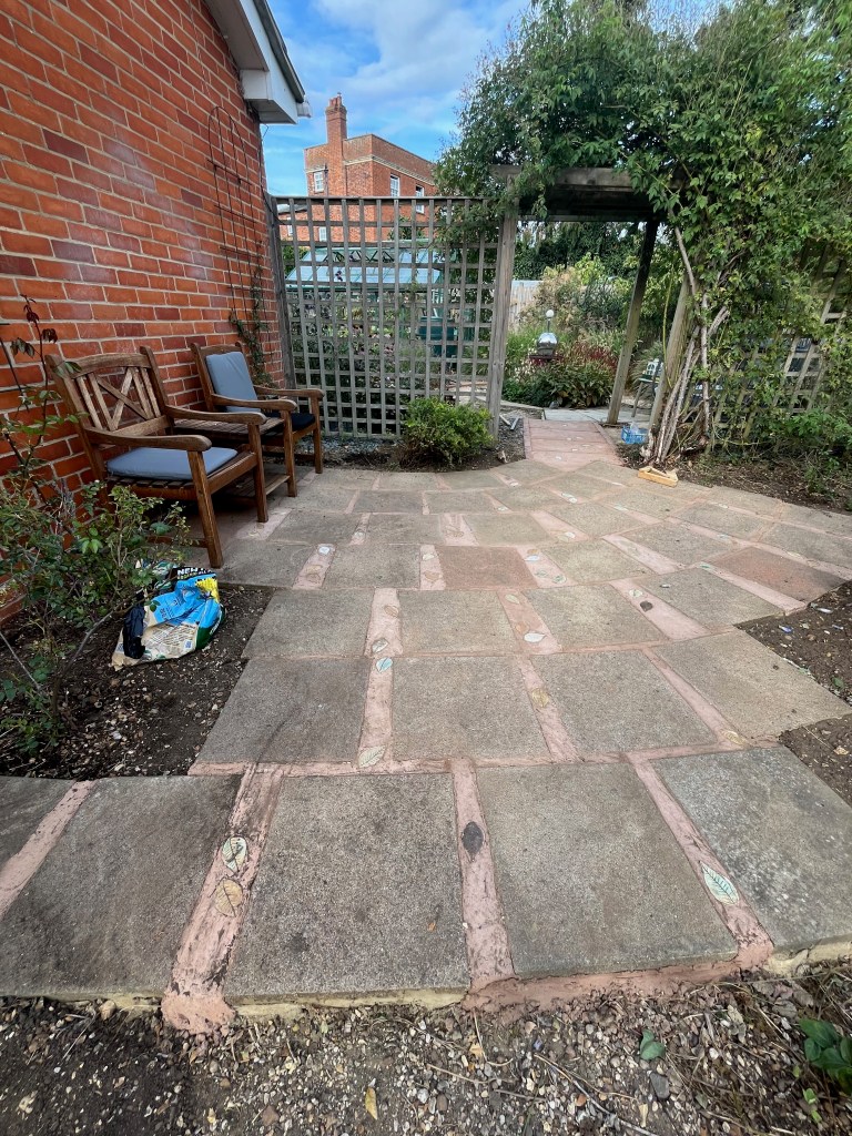 A landscaped garden area featuring a stone-paved path with pink detailing, wooden chairs, and a trellis covered with greenery, set against a brick wall and a clear sky.
