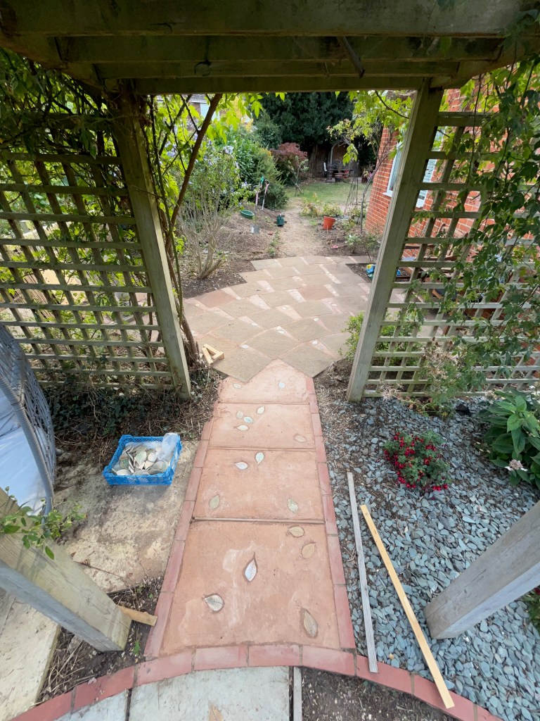 A garden pathway leading through a trellis, showcasing newly laid pavers with decorative leaves and surrounded by greenery.