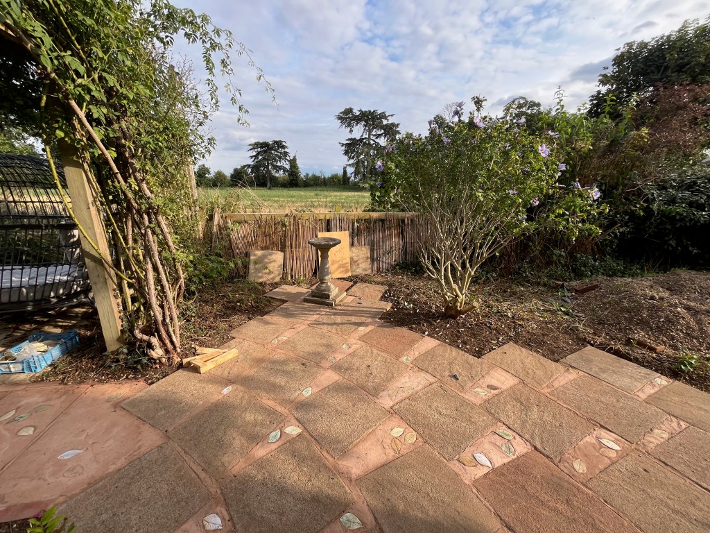 A renovated garden view featuring a circular stone patio, a bird bath, and lush greenery, with a tranquil rural background.
