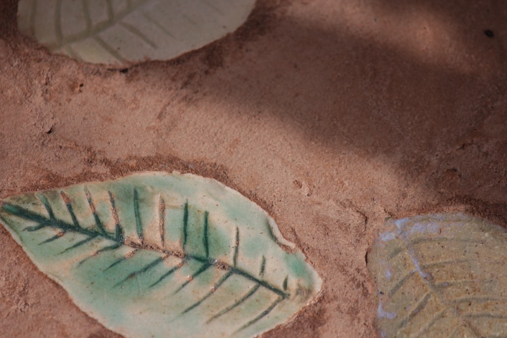 Close-up of leaf-shaped ceramic tiles embedded in a sandy-colored surface, showcasing intricate green details and textures.