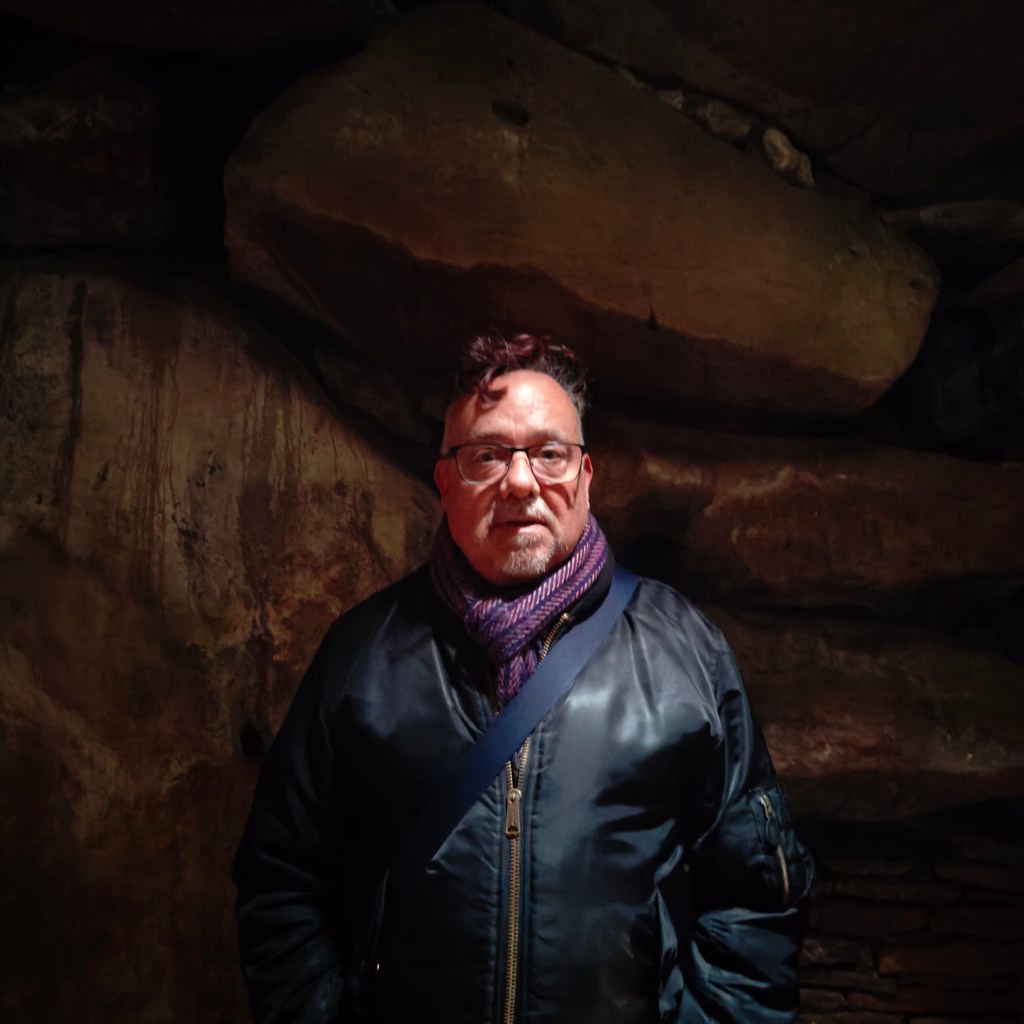 A person standing in a dimly lit chamber of West Kennet Long Barrow, surrounded by large stones and ancient rock formations.