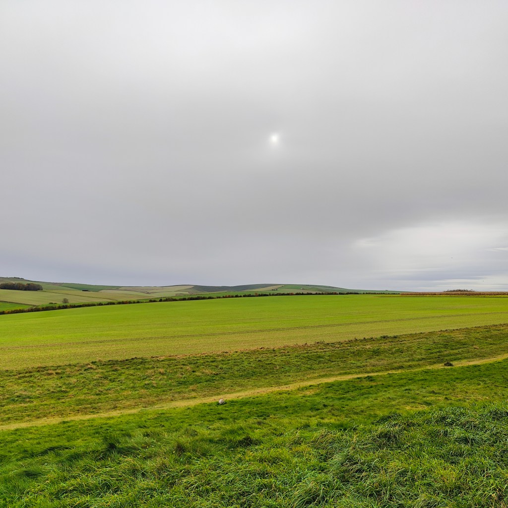 A panoramic view of rolling green fields under a cloudy sky, with a faint sun visible in the distance. The landscape features gentle hills and an open horizon.